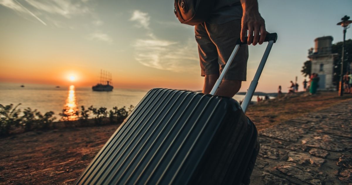 Voyageur tirant une valise rigide noire au coucher de soleil sur un quai bord de mer, soleil orangé en arrière-plan, préparation d'un combiné Maurice-Réunion